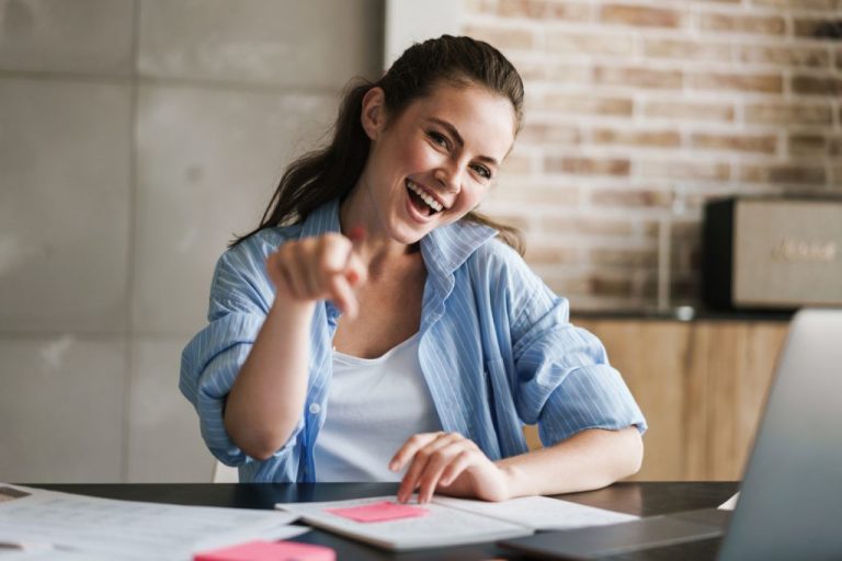 Egy kék inget viselő nő ül az íróasztalnál, papírokkal, mosolyogva, vidáman a kamera felé mutatva talán egy jól működő weboldal sikerét jelezve.